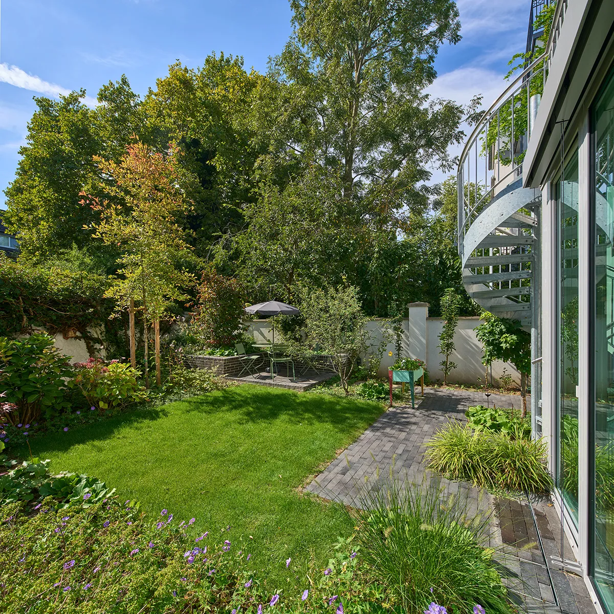 Gartenansicht der Maisonettewohnung mit Metall-Wendeltreppe, großzügiger Rasenfläche und Sitzplatz im hinteren Bereich.